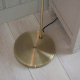 Endon Wentworth floor lamp base in antique brass, showing the circular weighted plate and slim stem on pale wooden floorboards beside a grey cabinet.