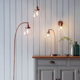 Endon Wentworth copper table lamp with clear glass shade and adjustable head, shown on an oak sideboard alongside coordinating pendant and wall lights.