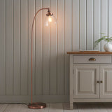 Endon Wentworth arched floor lamp in copper with clear glass dome shade and exposed E27 bulb, beside a cream sideboard against panelled walls.