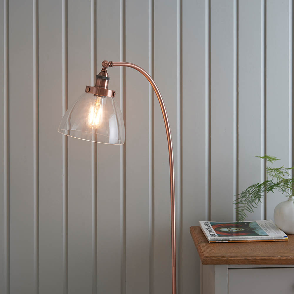 Endon Wentworth floor lamp in copper with an arched stem, clear glass shade and lit E27 bulb, beside a light grey panelled wall and oak side table.