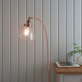 Endon Wentworth floor lamp in copper with an arched stem, clear glass shade and lit E27 bulb, beside a light grey panelled wall and oak side table.