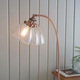 Endon Wentworth floor lamp in copper with arched stem, adjustable knuckle, clear glass shade and visible filament bulb, set against light panelled wall.