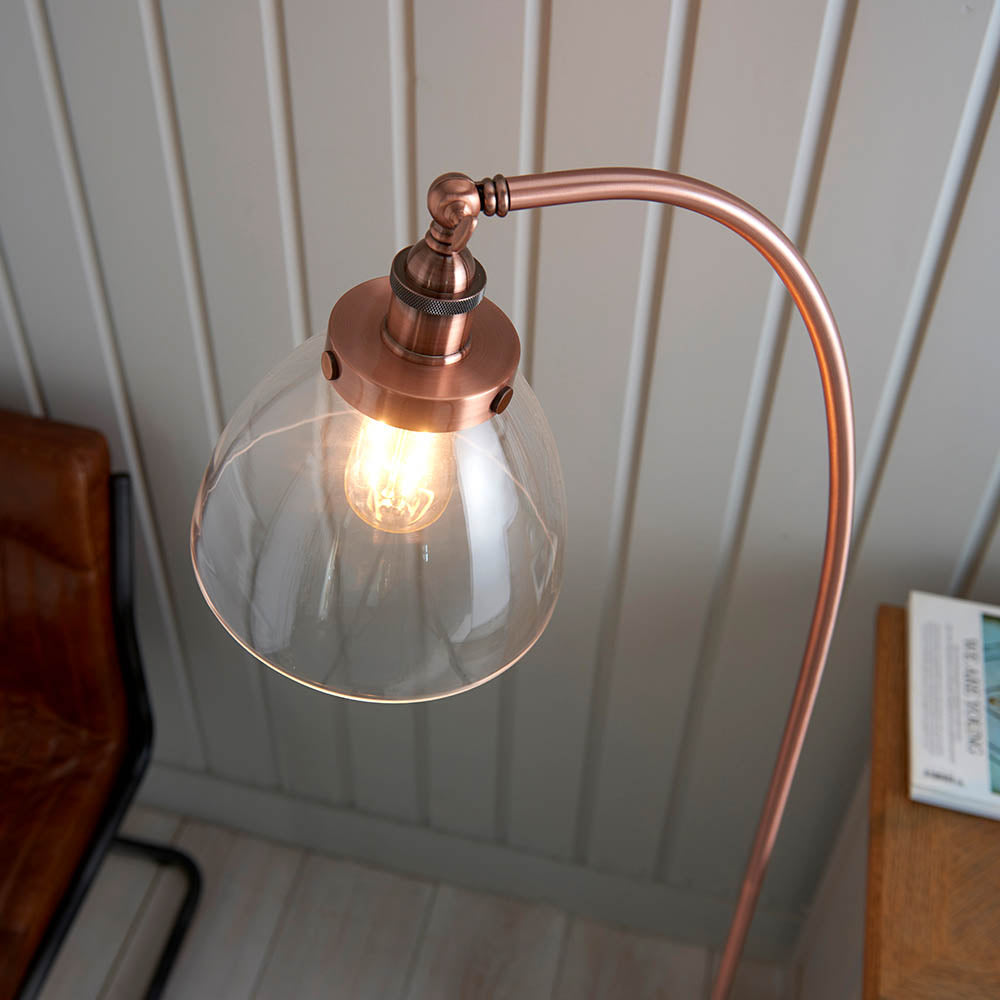 Endon Wentworth copper floor lamp with arched arm, knurled lamp holder, and clear glass shade housing an E27 bulb, shown beside a leather chair.