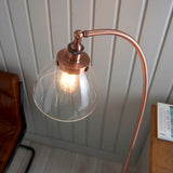 Endon Wentworth copper floor lamp with arched arm, knurled lamp holder, and clear glass shade housing an E27 bulb, shown beside a leather chair.