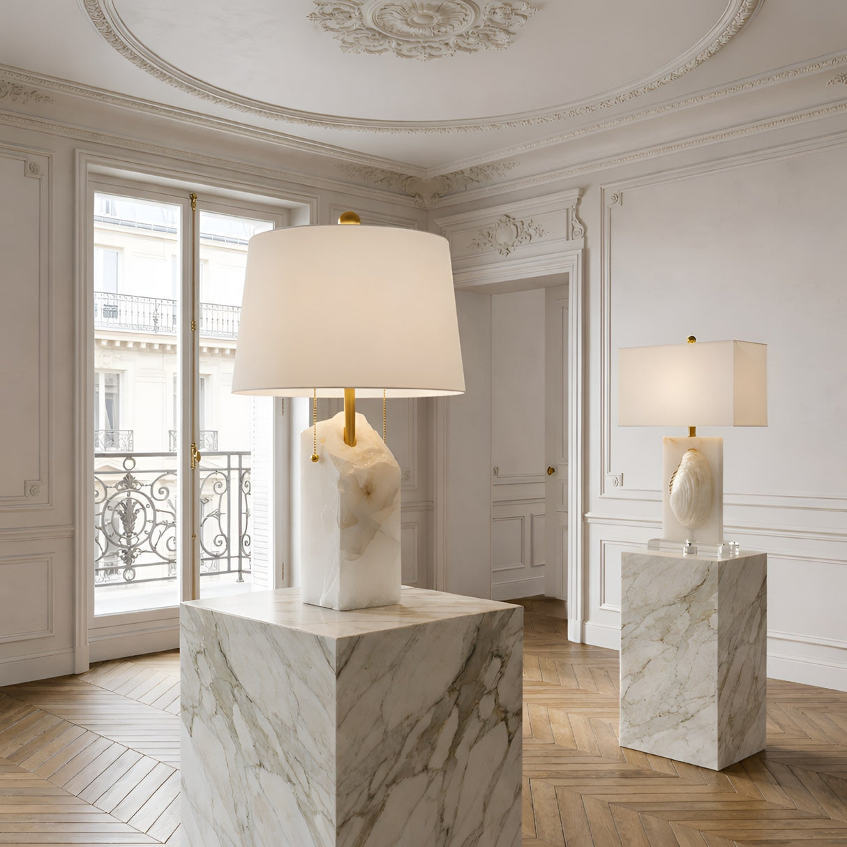 A bright, elegant room with ornate ceiling moldings, herringbone wood floors, and two Carved 1 Light Alabaster Table Lamps in brass & soft white atop marble pedestals. Large windows flood the space with natural light.