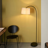 Endon Bevbourne arched floor lamp in matt black with tilting light grey fabric shade, beside a black chair and desk against a yellow wall.