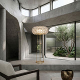 Briller LED Floor Lamp by Niori with clear acrylic sphere shade on a slim gold frame, in a concrete interior beside a boucle armchair and courtyard.