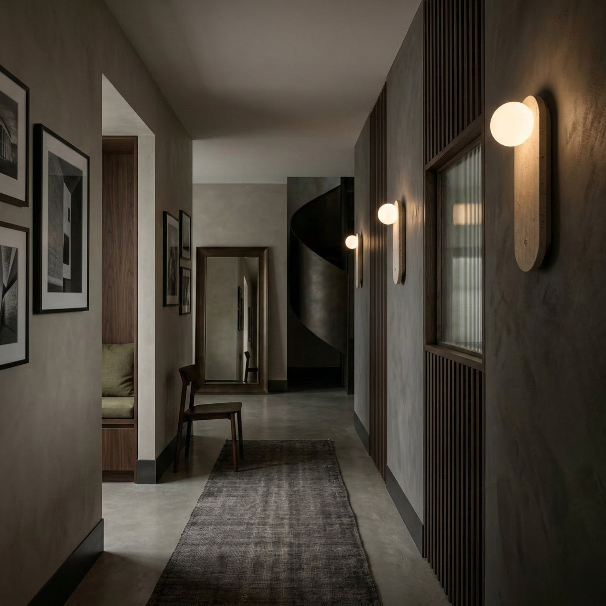A modern hallway with gray walls, art, a dark rug, a wooden chair by a tall mirror, and a spiral staircase features elegant Arvella 1 Light Travertine & Glass Wall Lights in light beige for sleek, sophisticated illumination.