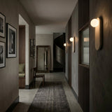 A modern hallway with gray walls, art, a dark rug, a wooden chair by a tall mirror, and a spiral staircase features elegant Arvella 1 Light Travertine & Glass Wall Lights in light beige for sleek, sophisticated illumination.