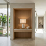 A bright, minimalist hallway features a built-in wooden niche displaying the Elvaris 1 Light Framed Marble Table Lamp in brass & white, alongside decorative objects and bowls. Large windows reveal lush greenery outside and fill the space with natural light.