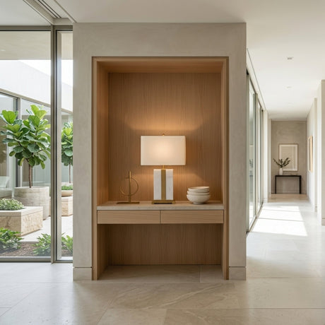 A bright, minimalist hallway features a built-in wooden niche displaying the Elvaris 1 Light Framed Marble Table Lamp in brass & white, alongside decorative objects and bowls. Large windows reveal lush greenery outside and fill the space with natural light.