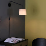 Endon Bevbourne plug-in wall light with matt black swing arm and light grey fabric shade, mounted above a dark desk with an open book.