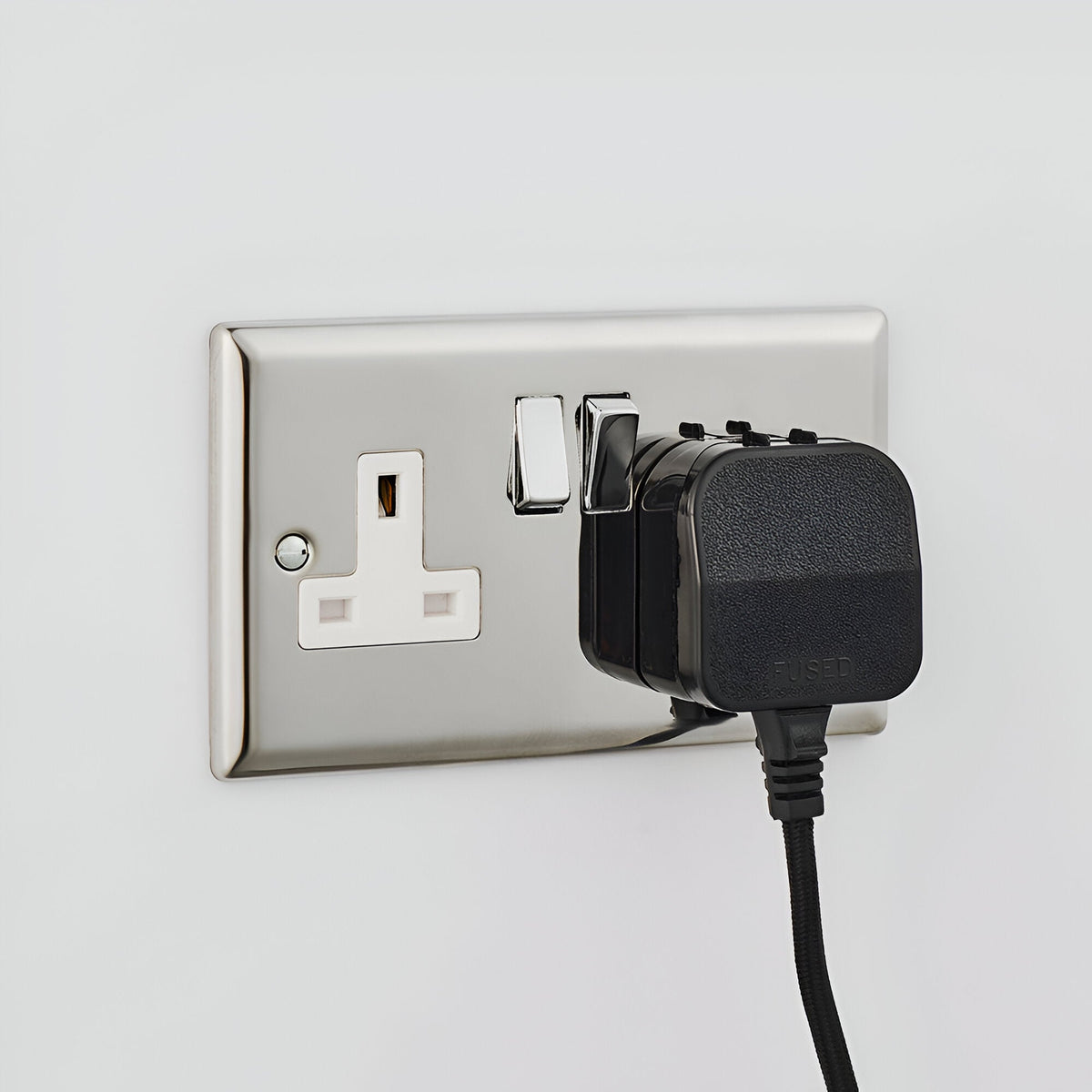 Black plug-in adapter with in-line fuse connected to a polished chrome UK wall socket, showing the Endon Bevbourne wall light's fabric flex and switch.