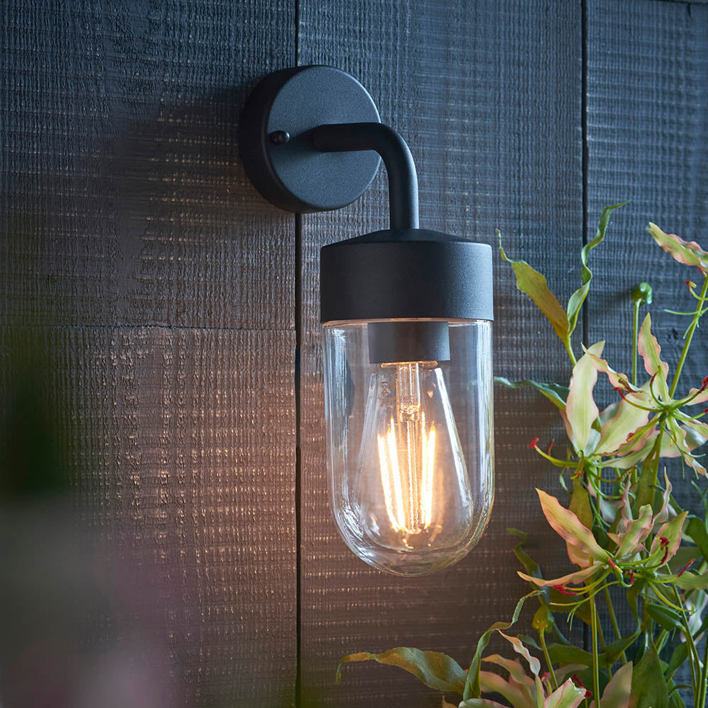 Endon Castfield IP44 outdoor wall light in matt black with clear glass cylinder shade and E27 filament bulb, mounted on dark timber cladding beside foliage.