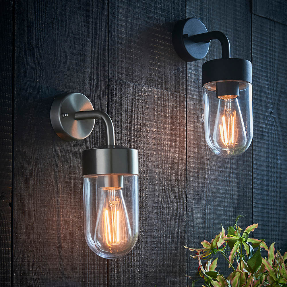 Endon Castfield outdoor wall lights in brushed steel and matt black, with clear glass cylinder casings and E27 filament bulbs, mounted on dark wood cladding.