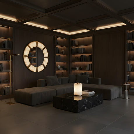 A modern, dimly lit living room with dark walls, built-in shelves, a large sectional, and a black marble coffee table features the Aurelior LED Small Ribbed Alabaster Table Lamp – Soft White glowing elegantly beneath a round backlit wall mirror.