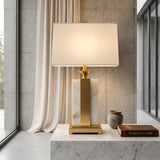 The Chandor 1 Light Alabaster Table Lamp in Brass & Soft White sits on a marble surface beside a brown book, The Brutalist Form, and a small ceramic vase, backdroped by a sheer curtain and concrete wall.