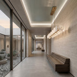 A modern hallway with beige stone walls, a sleek wooden bench, and large windows overlooking a minimalist courtyard. At the ceiling hangs the Solaryn LED Linear Disc Alabaster Suspension Light in soft white & brass. Art pieces are displayed at the hall’s end.