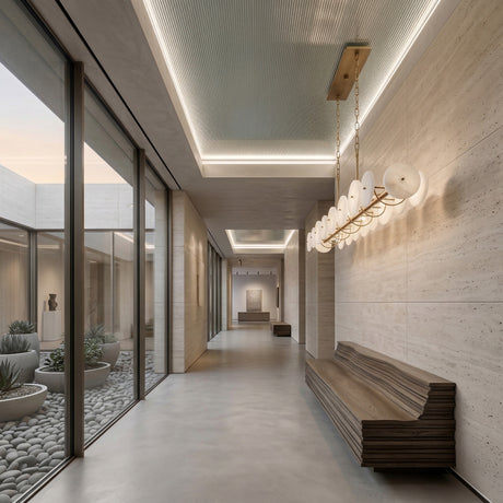 A modern hallway with beige stone walls, a sleek wooden bench, and large windows overlooking a minimalist courtyard. At the ceiling hangs the Solaryn LED Linear Disc Alabaster Suspension Light in soft white & brass. Art pieces are displayed at the hall’s end.