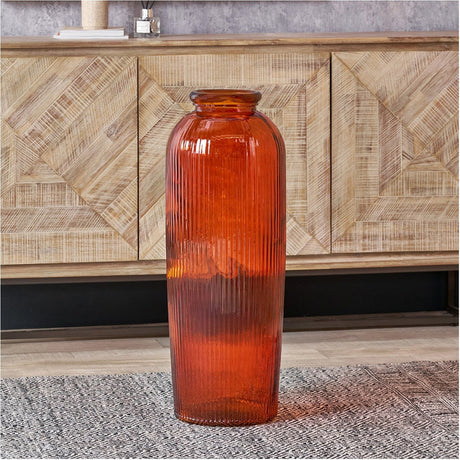 Tall orange recycled glass ribbed vase with narrow neck, standing on a patterned rug in front of a wood-panelled sideboard in a living room setting.