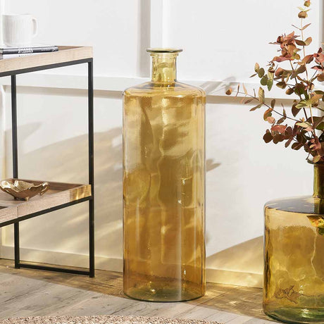 Amber Recycled Tall Glass Bottle Vase, 75cm, with a narrow neck and cylindrical body, styled on a wooden floor beside a console table and a smaller green glass vase with foliage.