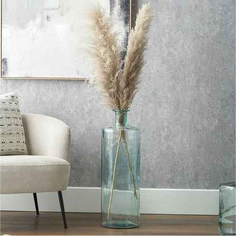 Clear recycled glass tall bottle vase, 75cm, holding pampas grass stems, standing on a wooden floor beside an armchair in a living room.