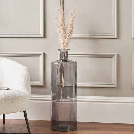 Grey recycled glass bottle vase, 75cm tall, cylindrical form with dried pampas grass stems, shown beside a cream boucle chair in a living room with panelled walls.