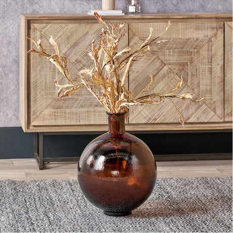 Round pecan brown recycled glass vase, 34cm wide, holding golden dried branch stems, styled on a rug in front of a wooden sideboard.