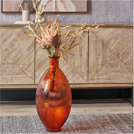 Tall orange recycled ribbed glass vase with dried floral stems, styled on a rug in front of a wooden sideboard in a living room setting.