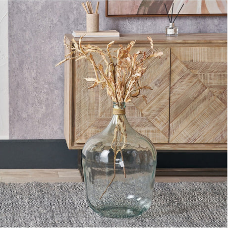 Clear recycled glass large bottle vase, 56cm, with a raffia-wrapped neck, holding dried foliage on the floor beside a wooden sideboard.