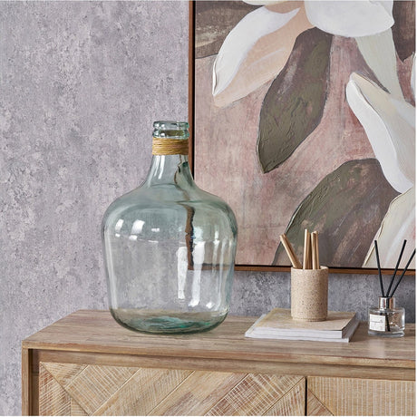 Clear recycled glass demijohn-style bottle vase, 42cm, with raffia-wrapped neck, shown on a wooden sideboard beside books and a reed diffuser.