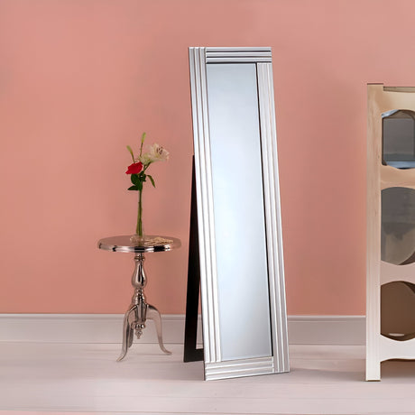 The Edge Free Standing Mirror - 150x40cm, with its sleek design, leans elegantly against the pink wall. Beside it, a minimalist metallic side table supports a vase filled with red and white roses. A glimpse of a light-colored cabinet is visible on the right.