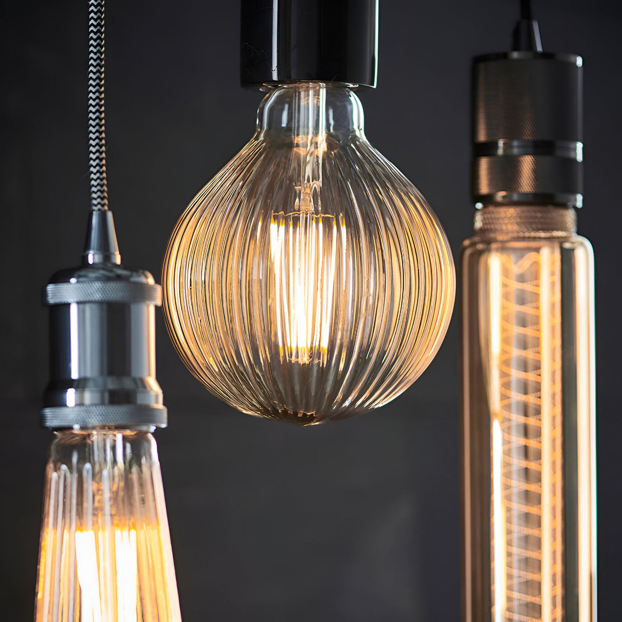 Ribbed globe LED filament bulb with amber tinted glass and E27 screw cap, shown lit alongside other pendant fittings against a dark background.
