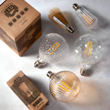 Assorted LED filament bulbs including a ribbed amber globe with E27 screw cap, alongside clear ST64 and geometric-cut globe bulbs with kraft packaging.