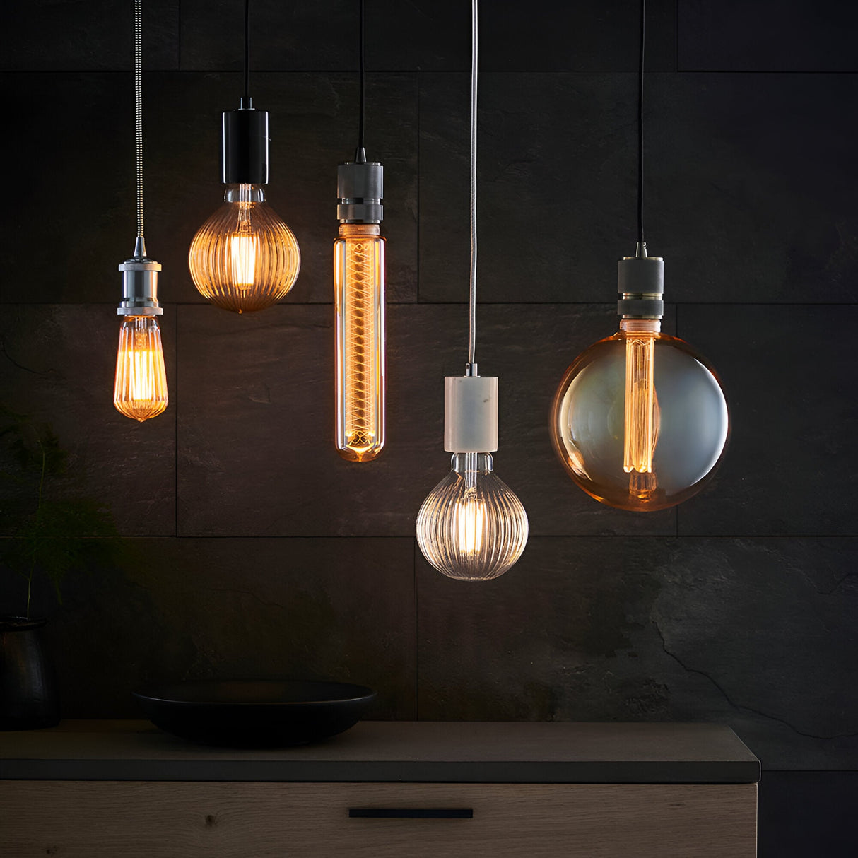 Assorted pendant lights with ribbed and smooth glass shades, including a clear E27 LED filament ribbed globe bulb, in a dark-toned interior setting.