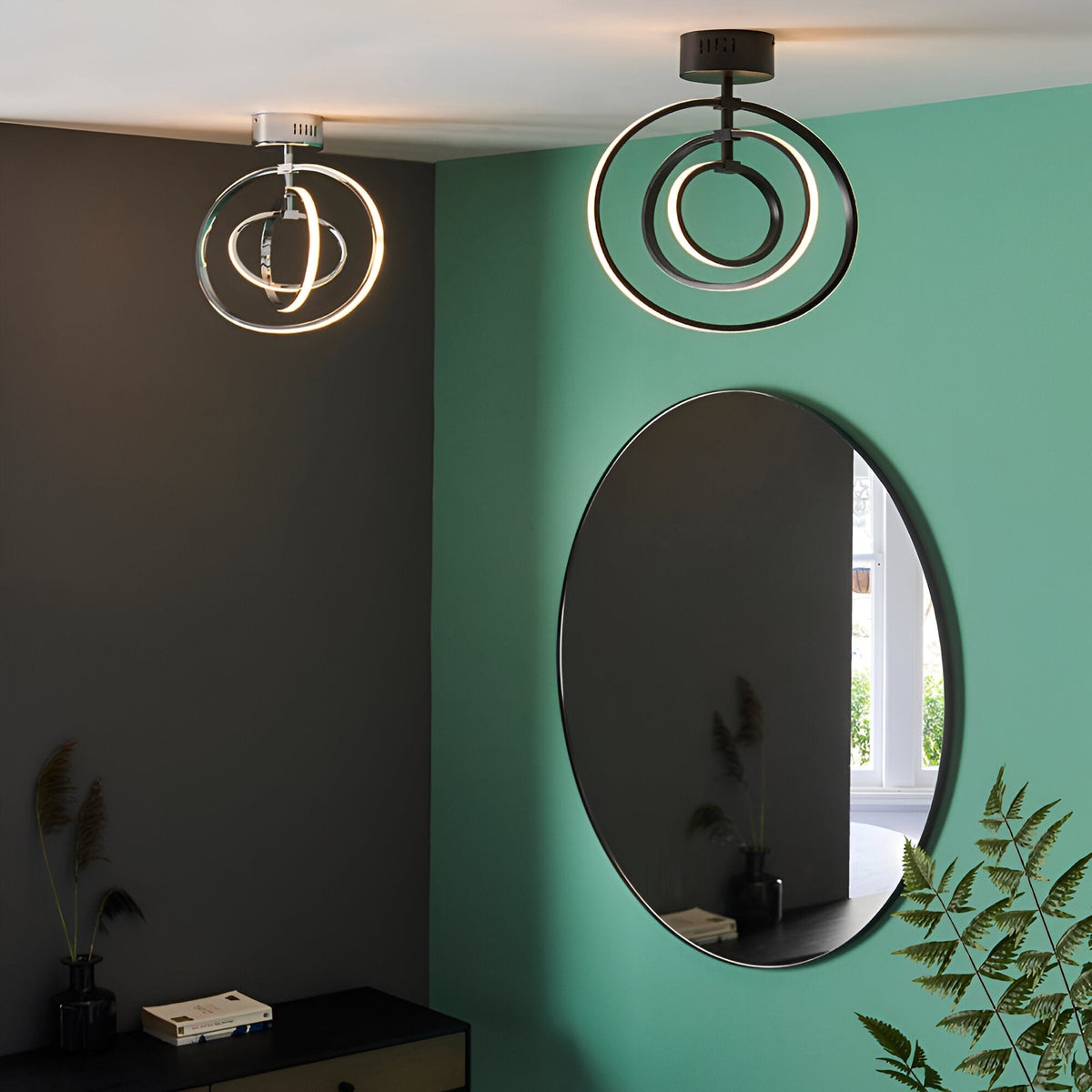 Endon Corso matt black semi-flush LED ceiling light with concentric rings, shown beside a chrome version above an oval mirror on a green wall.
