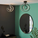 Endon Corso matt black semi-flush LED ceiling light with concentric rings, shown beside a chrome version above an oval mirror on a green wall.