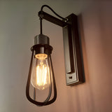 Endon Camden industrial wall light with caged teardrop shade, exposed E27 filament bulb, and chrome and black stepped bracket mounted on a pale wall.