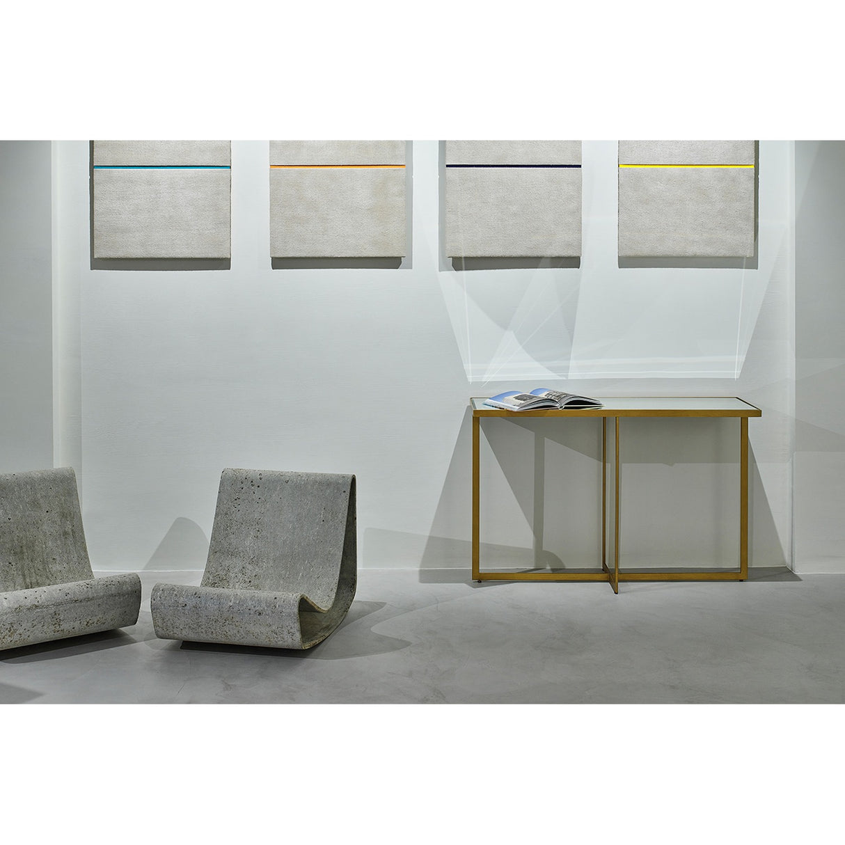 Tablo Bronze Mirrored Glass Console Table with crossed X-frame legs and mirrored top, shown against a gallery wall with framed art and two sculptural concrete chairs.