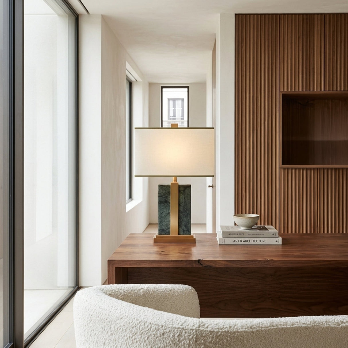 A modern office showcases a wooden desk with the Elvaris 1 Light Framed Marble Table Lamp in brass & green, a stack of books, and a bowl. The space includes a textured wood accent wall, white armchair, and floor-to-ceiling windows.