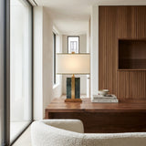 A modern office showcases a wooden desk with the Elvaris 1 Light Framed Marble Table Lamp in brass & green, a stack of books, and a bowl. The space includes a textured wood accent wall, white armchair, and floor-to-ceiling windows.
