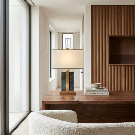 A modern office showcases a wooden desk with the Elvaris 1 Light Framed Marble Table Lamp in brass & green, a stack of books, and a bowl. The space includes a textured wood accent wall, white armchair, and floor-to-ceiling windows.
