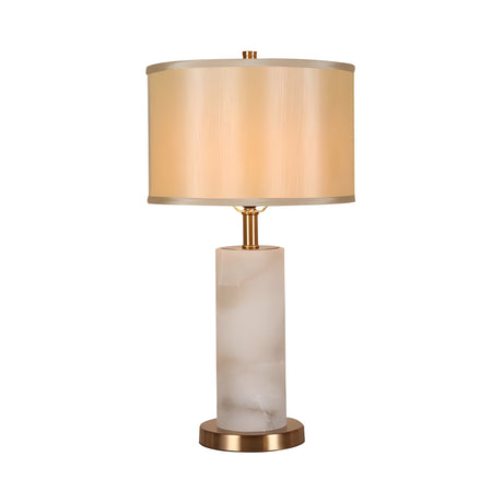 The Calynor 1 Light Column Alabaster Table Lamp features a cylindrical frosted white Spanish alabaster base, brass round pedestal, and beige drum shade. Lamp is off and set against a plain white background.