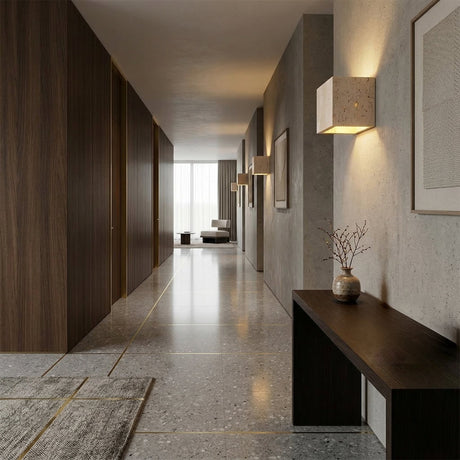 A modern hallway with stone tile floors, dark wood walls, Cavaro LED Cube Travertine Wall Lights in light beige, a slim console table with a vase, framed artwork, and a distant seating area by large windows with sheer curtains.
