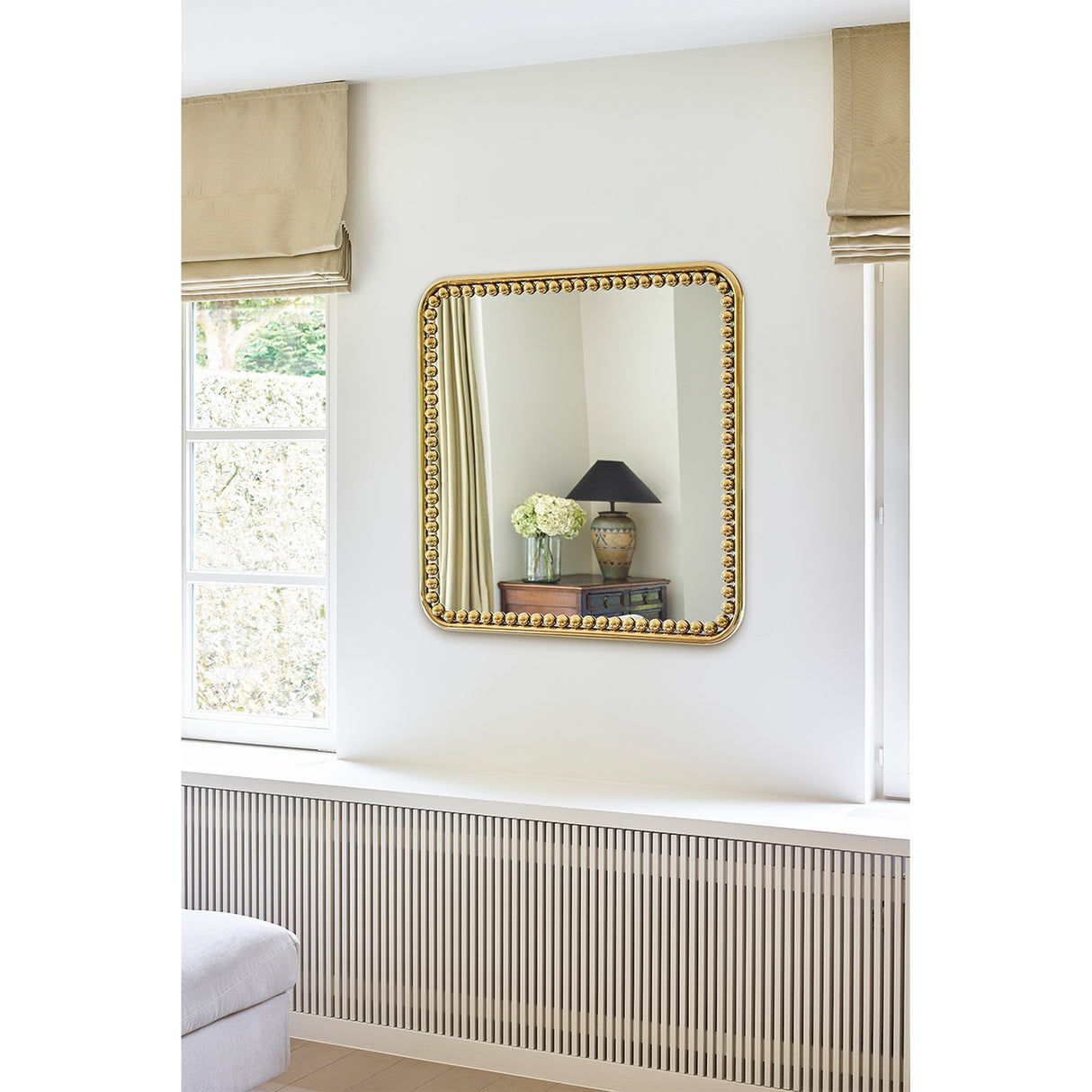 Diva Beaded Gold Frame Square Wall Mirror 95cm on a light bedroom wall, reflecting a lamp and side table, with a beaded gold metal frame around the glass.
