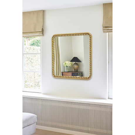 Diva Beaded Gold Frame Square Wall Mirror 95cm on a light bedroom wall, reflecting a lamp and side table, with a beaded gold metal frame around the glass.