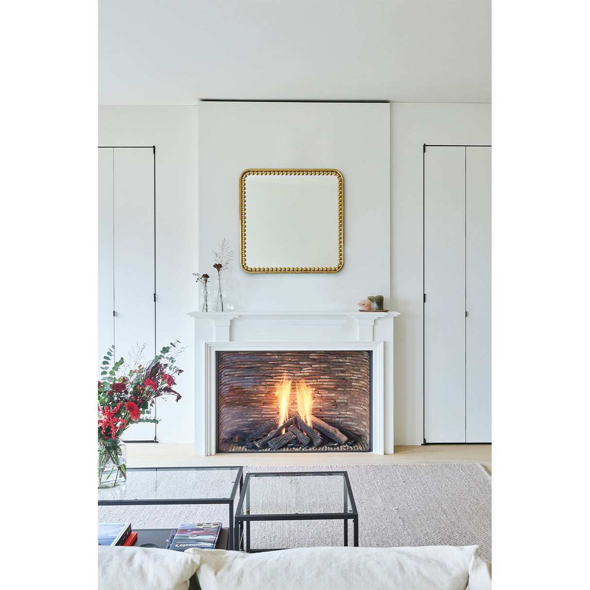 Diva Square 95cm gold beaded-frame wall mirror hung above a white fireplace mantel in a living room, with a lit fire, mirror surface reflecting the room.