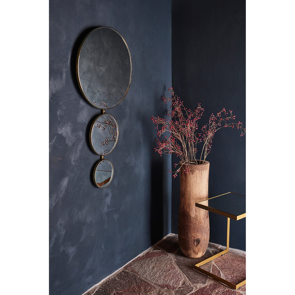 Opera Three Circle Gold Rimmed Wall Mirror 119cm hung on a dark wall, three joined round mirrors in decreasing size with slim gold rims, above a wooden vase with red berry stems.