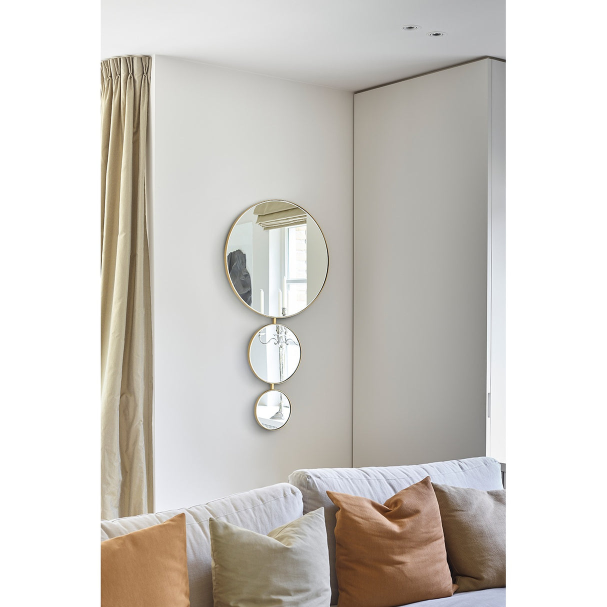 Opera Three Circle Gold Rimmed Wall Mirror 119cm featuring three joined circular mirrors in decreasing size, mounted on a light living room wall above a sofa with cushions.