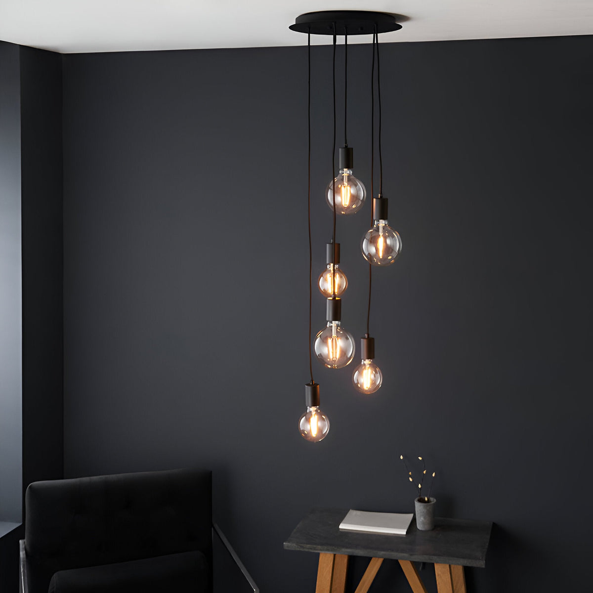 Marlowe 6 Light Pendant in matt black metal, six exposed bulbs on staggered black fabric flex drops, shown against a dark wall above a side table.